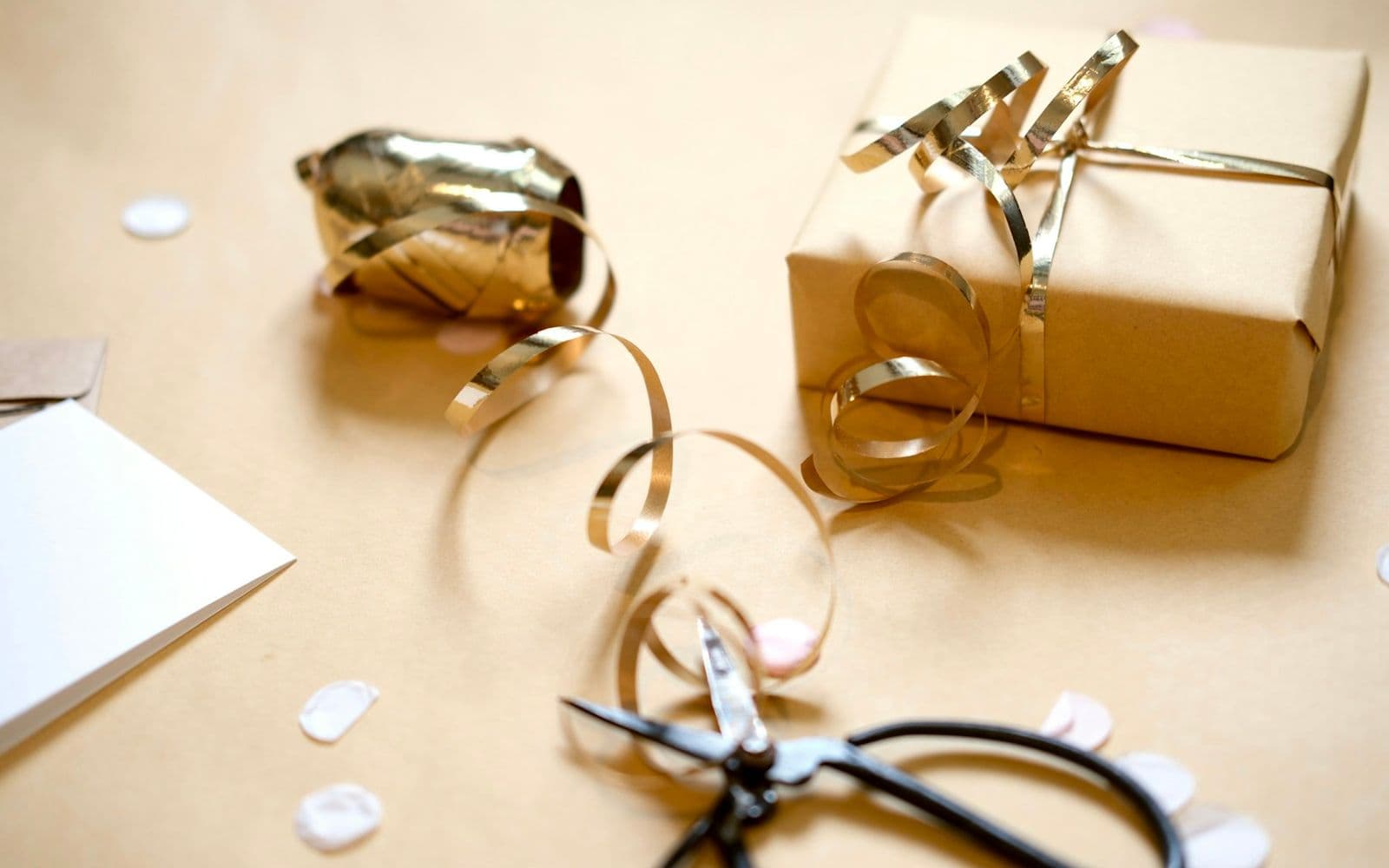 Kraft-paper wrapped gift tied with gold curling ribbon beside scissors, a white card and paper confetti