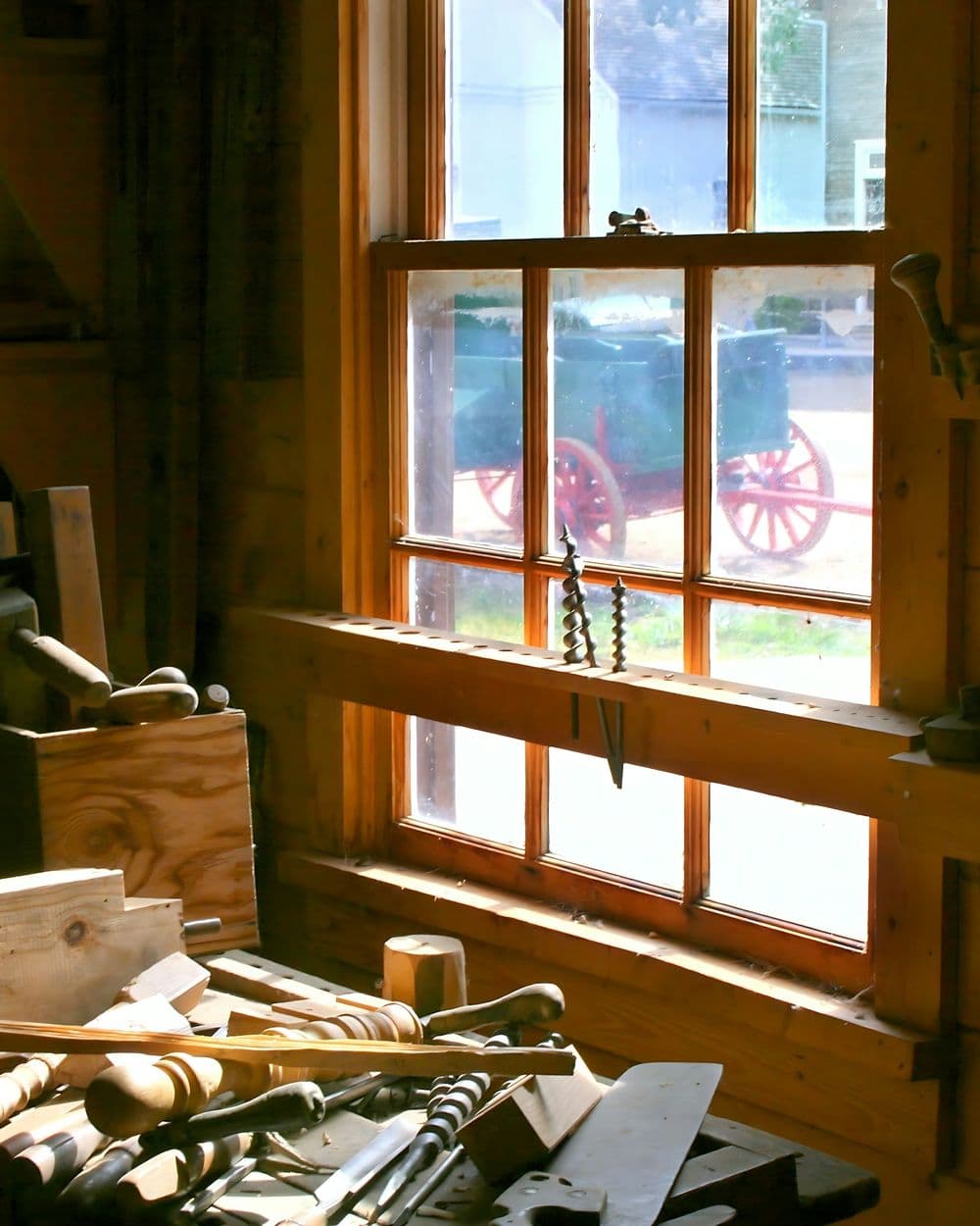 Sunlit woodworking shop window above a workbench crowded with planes, chisels and wooden mallets