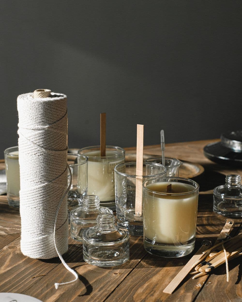 Candle-making supplies: cotton wick spool, wooden wicks and glass jars of wax on a table