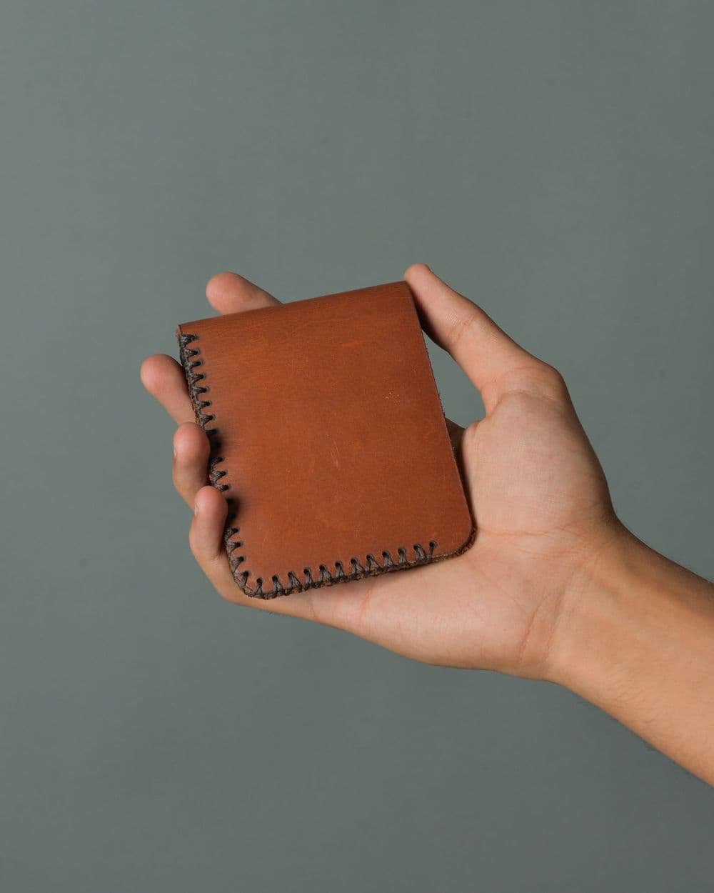 Hand holding a small brown stitched leather wallet against a grey wall