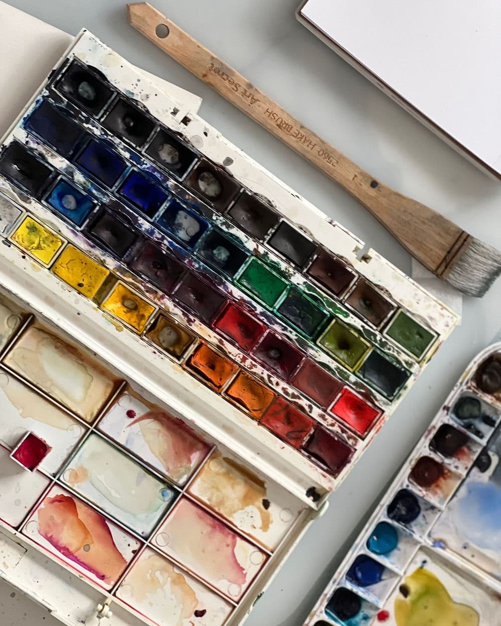 Well-used watercolor pan set with mixing wells beside a flat paintbrush