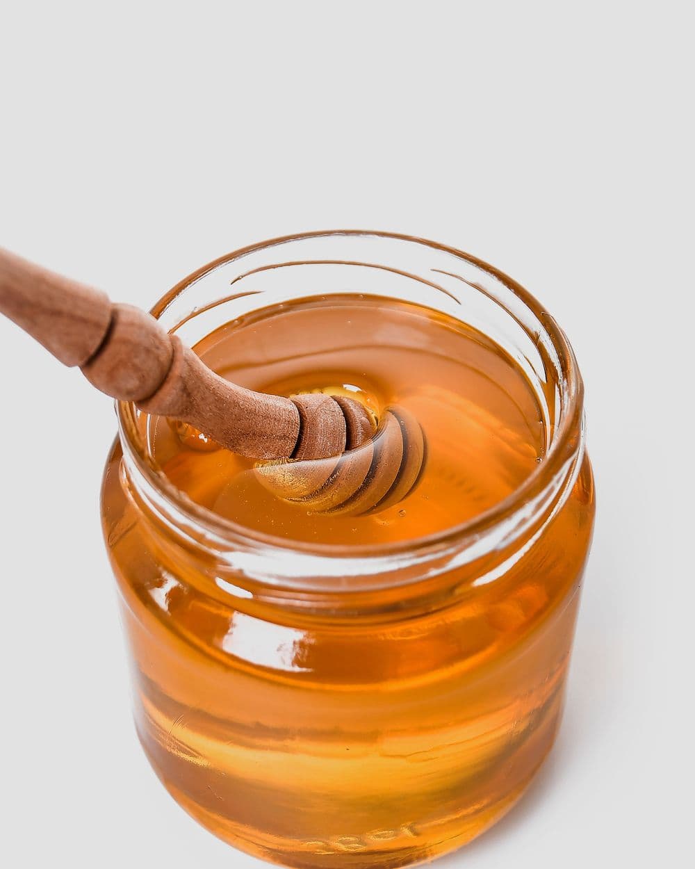 Glass jar of golden honey with a wooden honey dipper on a white background