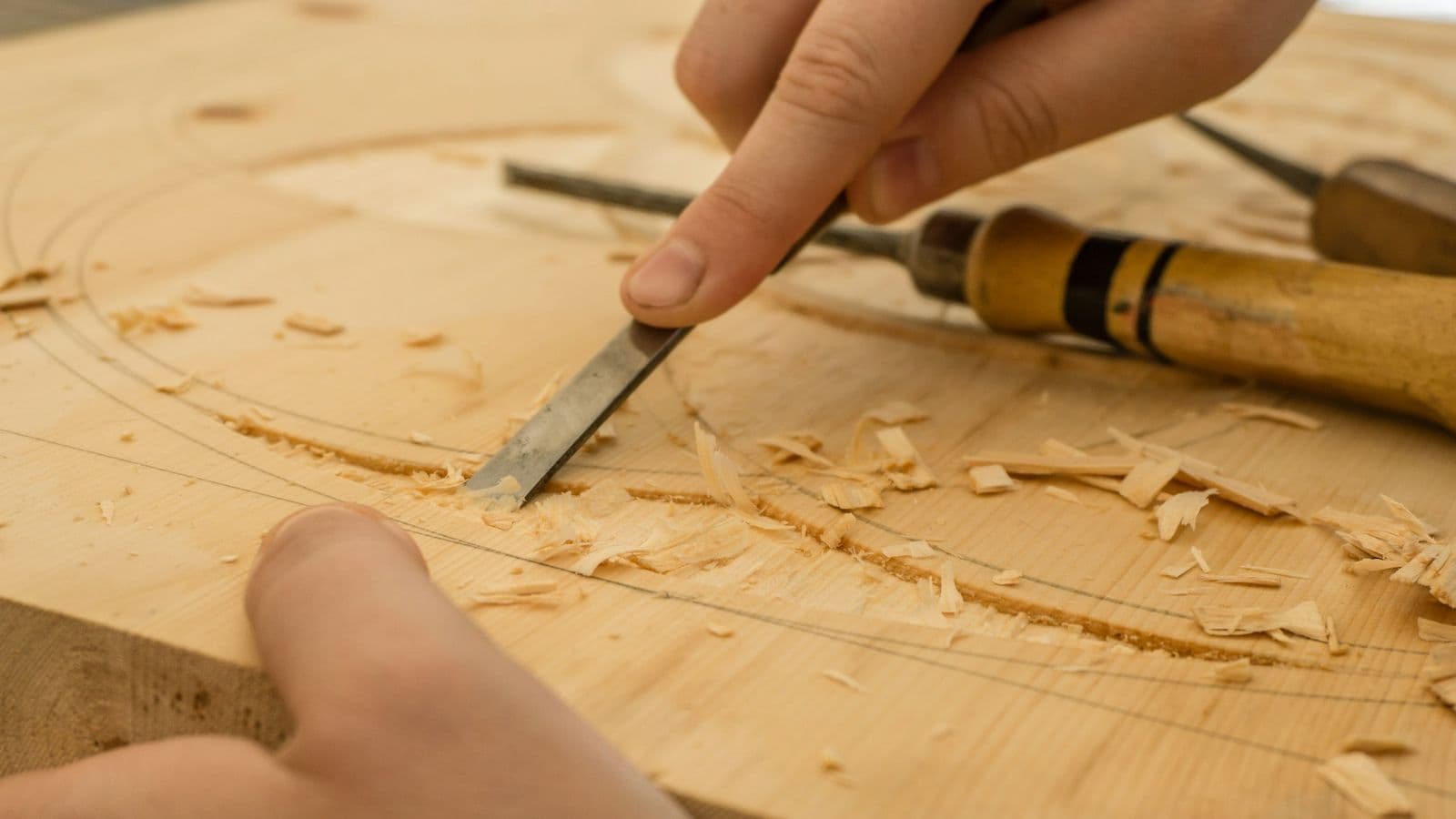 Hands guiding a wood chisel through a pale wooden board, with curled shavings along a pencil-marked arc
