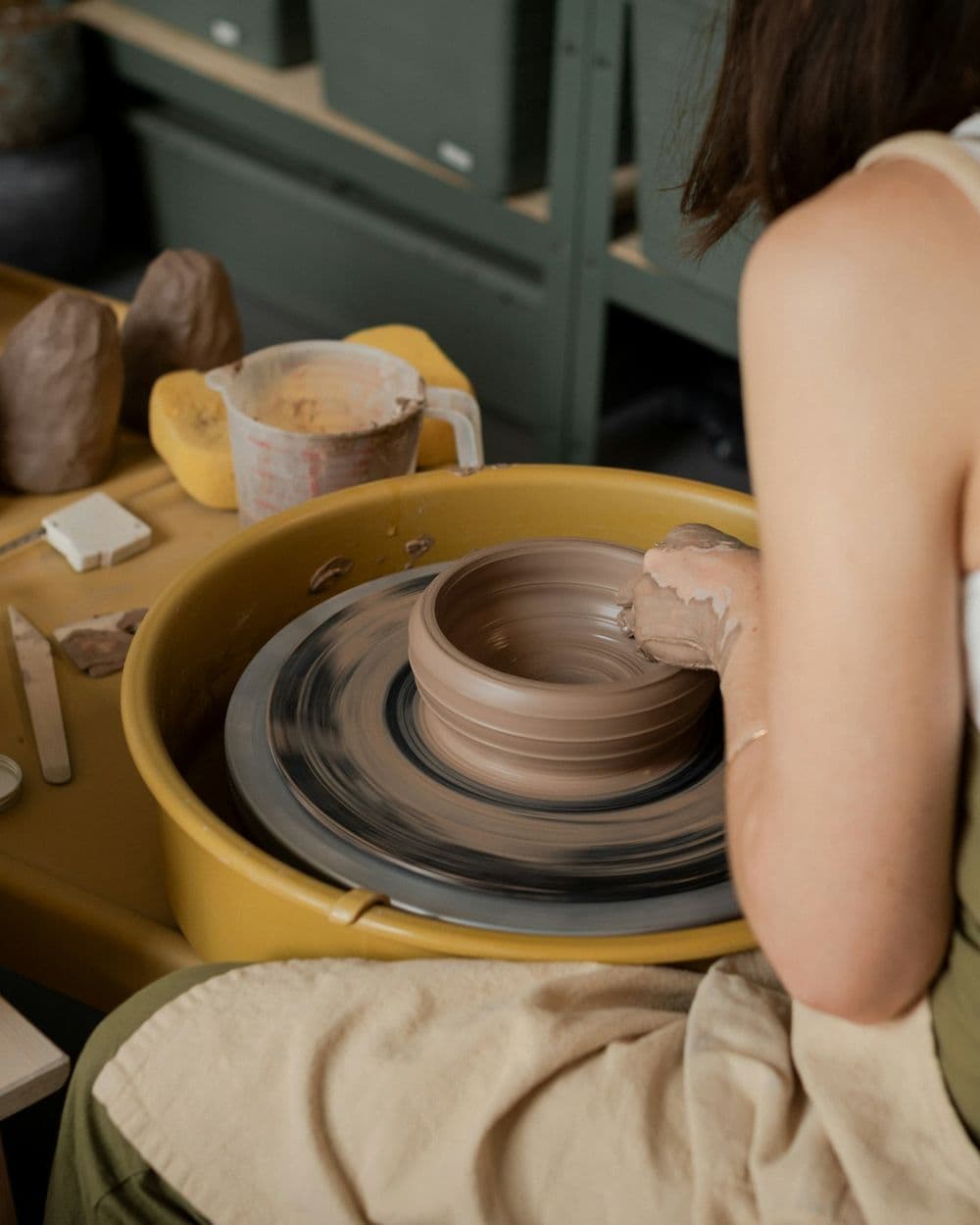 Potter at a wheel shaping a clay bowl, with tools, sponges and a water jug on the bench