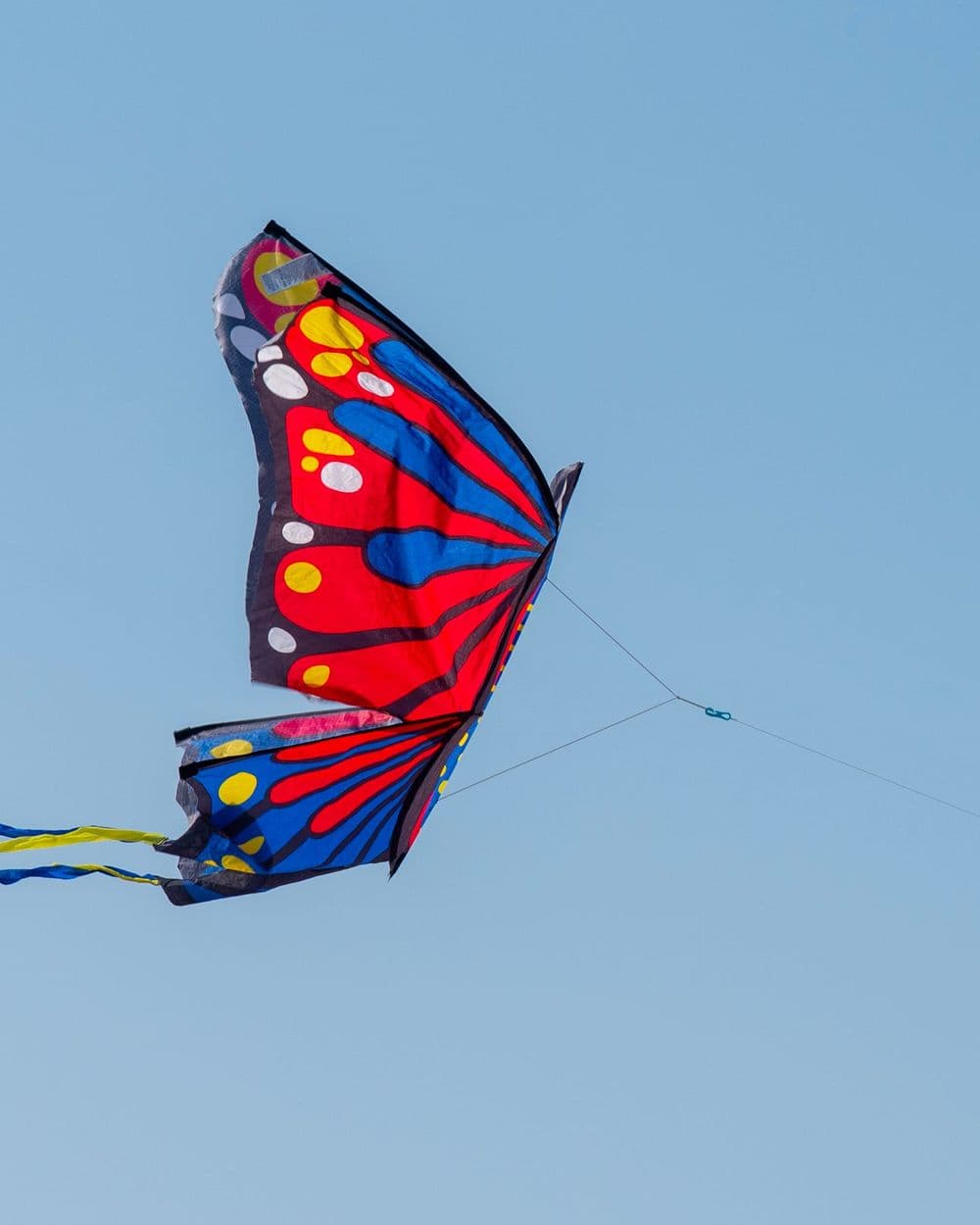 Red, blue and yellow butterfly kite with ribbon tails flying in a clear blue sky